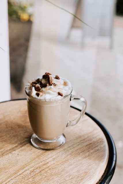 A Calhoun Coffee latte topped with whipped cream and chopped pecans in a footed glass mug on a wooden patio table.