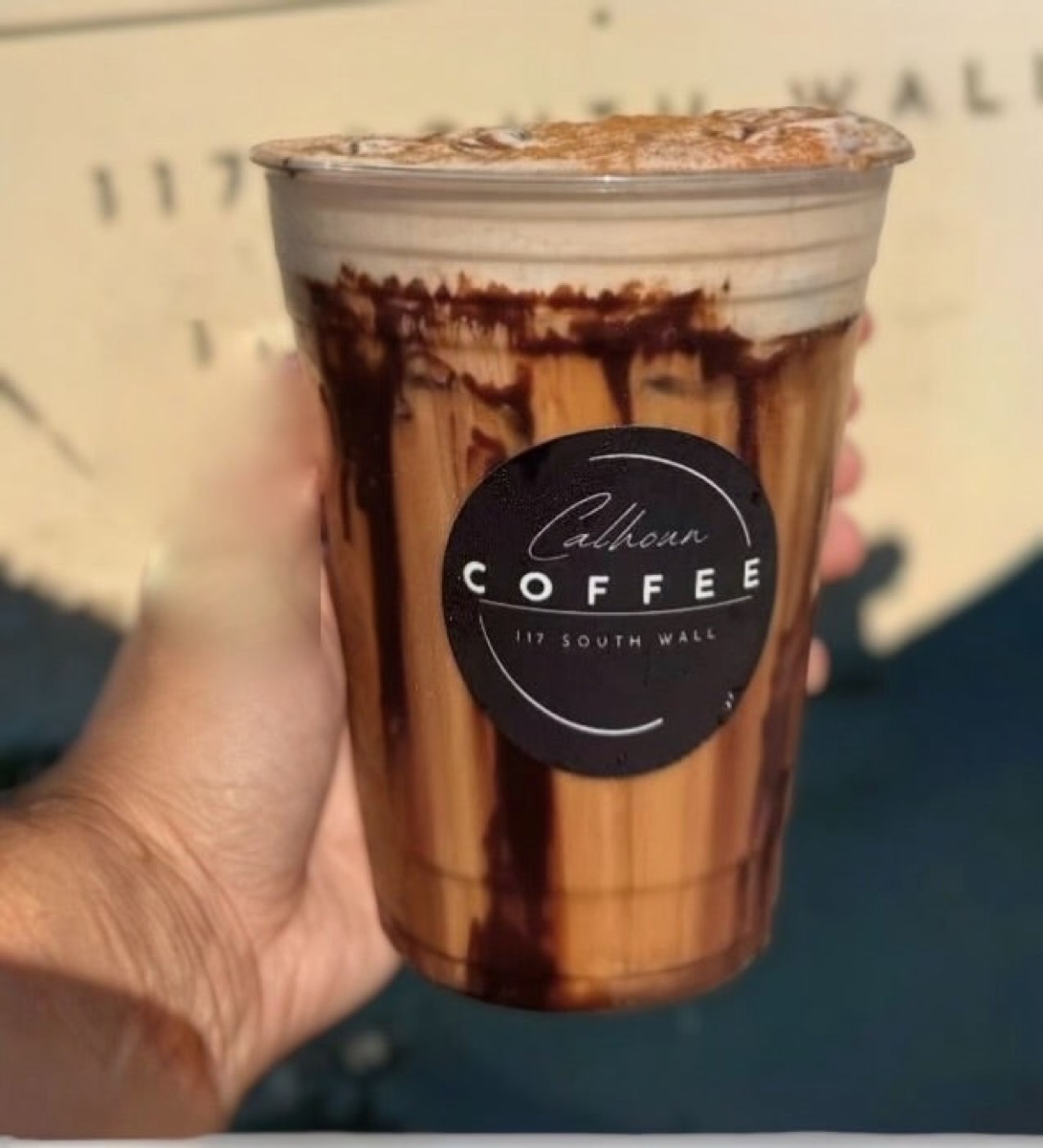An iced Calhoun Coffee specialty drink in a branded cup, held in hand — every order made fresh.